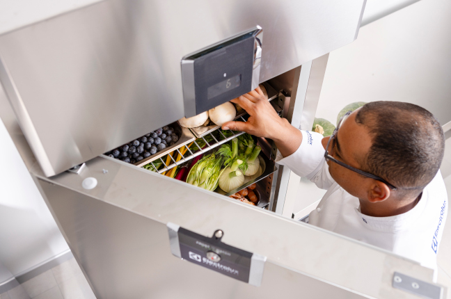 Chef reaching into a commercial refrigerator filled with fresh vegetables and fruits, including blueberries, mushrooms, lettuce, and carrots.