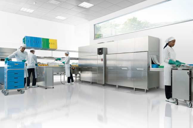In the middle of a pristine white room, a group of people wearing sterile white coats, hairnets, and turquoise gloves are operating professional dishwashing machinery. The room is modern and bright with large windows. The equipment is made of stainless steel and includes large washing machines and a series of conveyors. On the left, two people are working together near a cart with blue containers. Another person on the right is pushing a similar cart. The overall scene suggests a high level of hygiene and an industrial or medical setting.