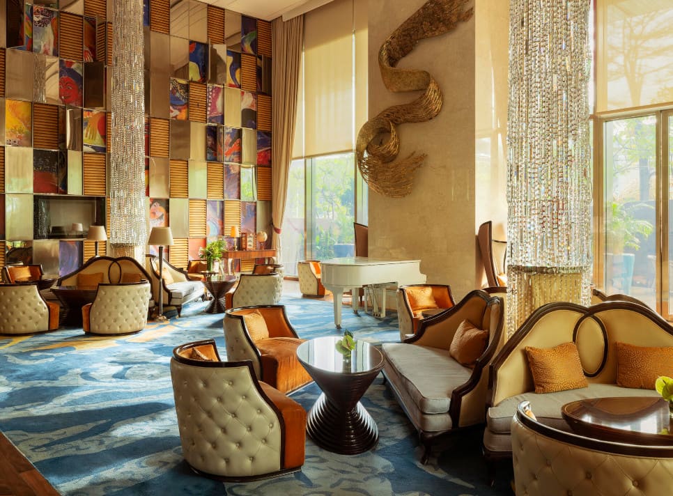 Luxurious hotel lounge with elegant furniture, a white grand piano, colorful wall art, and tall windows letting in natural light.